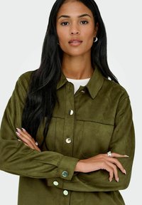 Olive green suede shirt with a collared neck, front pockets, and metallic button details; smooth texture; long sleeves with buttoned cuffs.