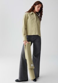 Lichtgroene button-up shirt met een voorzak, gecombineerd met hoge taille, brede donkergrijze jeans. Model houdt een geribbelde lichtgroene accessoire vast.