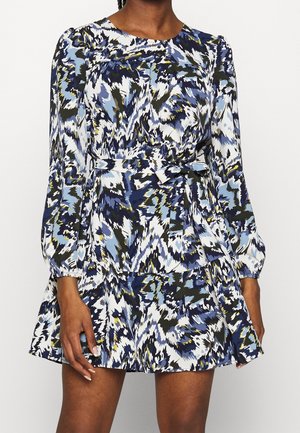 Woman wearing a belted, long-sleeve dress with a blue, white, black, and yellow abstract pattern and a flared skirt.