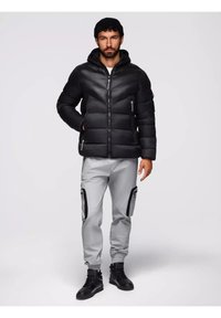 Giacca a vento nera con cappuccio, dotata di sezioni trapuntate e tasche con zip, indossata con pantaloni jogger grigi e stivali neri.