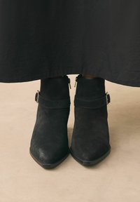 Botines de ante negros con punta afilada, cierre de cremallera y un detalle de correa alrededor de la parte superior. Ambientados en un fondo neutro.