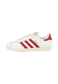 White sports shoe with red stripes and textured accents, featuring a round toe, lace-up design, and a textured rubber sole. Gold 'Gazelle' branding.