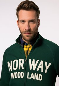Green zip-up sweatshirt featuring large white lettering "NOR WAY WOOD LAND." Worn over a yellow plaid shirt with a green and blue pattern.