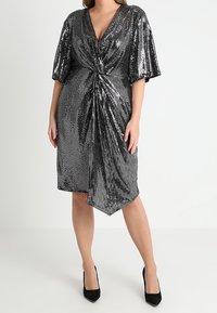 Robe noire à sequins avec un décolleté en V profond, manches courtes et détail torsadé à l'avant. Longueur genou avec une silhouette fluide, associée à des talons noirs.