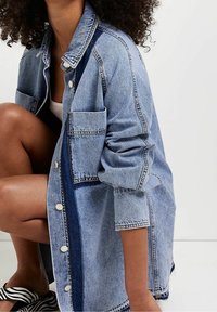 Person som knäböjer iförd en överdimensionerad ljusblå denimjacka med stora fickor och zebramönstrade sandaler mot en enkel bakgrund.