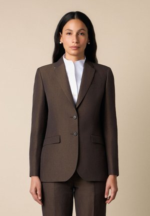 Kobieta o długich czarnych włosach, nosząca perłowe kolczyki, brązową marynarkę, dopasowane spodnie oraz białą koszulę z wysokim kołnierzem na beżowym tle.