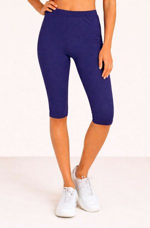 Femme portant des leggings bleu marine jusqu'aux genoux et des chaussures de sport blanches, debout avec une main sur la hanche sur un fond uni.