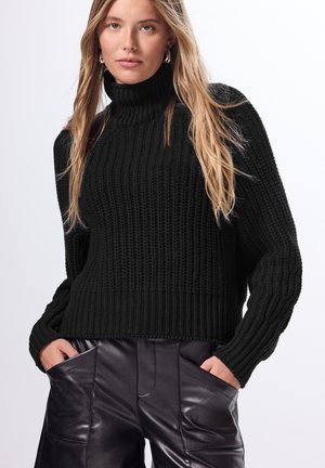 Maglione lavorato a maglia nero con colletto alto, texture a costine e maniche a palloncino, abbinato a pantaloni in pelle neri eleganti con tasche laterali.