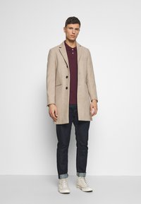 Beige wollen jas met een geknipt revers, drie knopen en zijzakken, gecombineerd met een bordeauxrood poloshirt en donkere jeans. Witte sneakers.