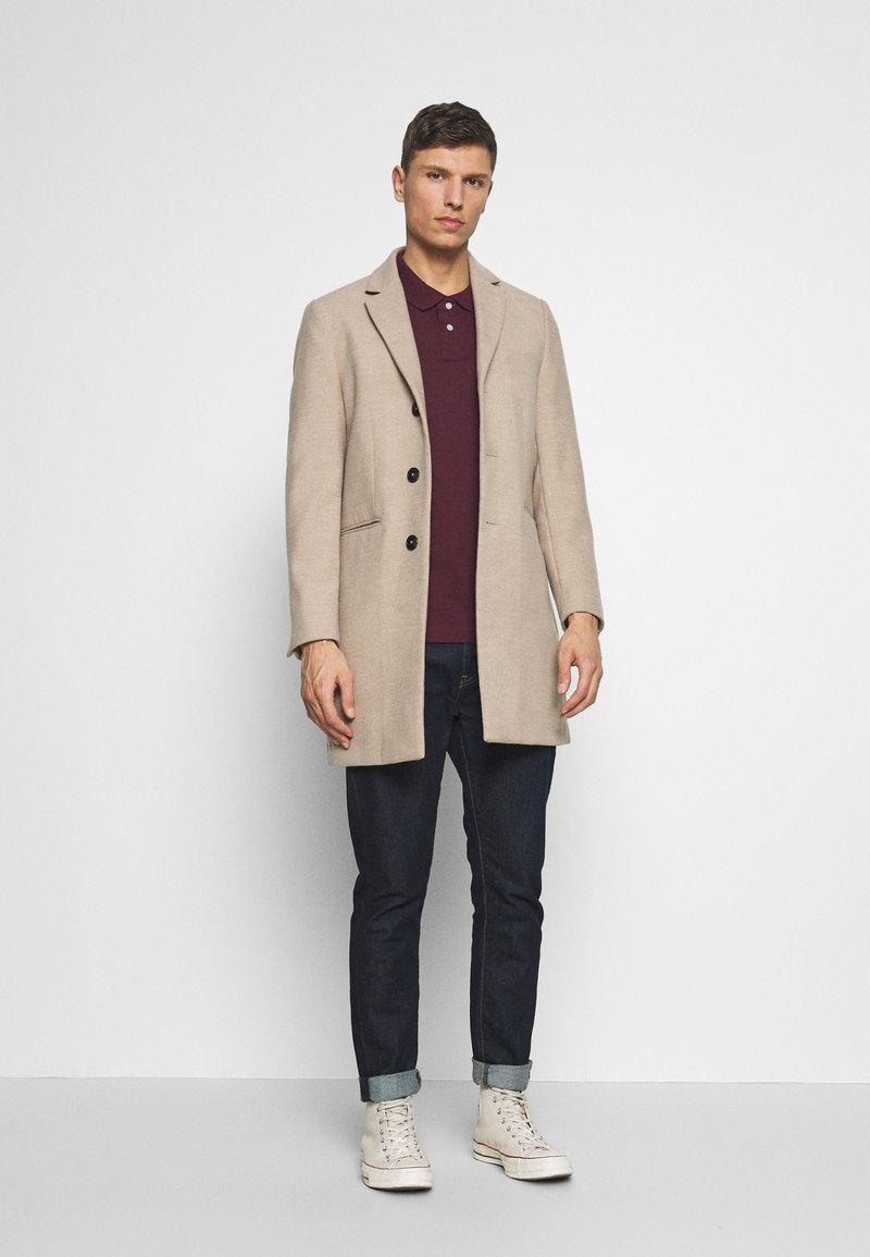 Beige wollen jas met een geknipt revers, drie knopen en zijzakken, gecombineerd met een bordeauxrood poloshirt en donkere jeans. Witte sneakers.