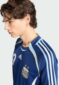 Giovane uomo con capelli ricci castani che indossa una maglia sportiva blu Adidas con il logo dell'Associazione Calcistica Argentina (AFA) e il numero 10.