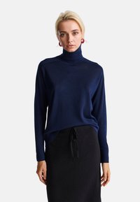 Maglione a collo alto blu navy con maniche lunghe, tessuto morbido e vestibilità rilassata, abbinato a una gonna nera con vita regolabile.