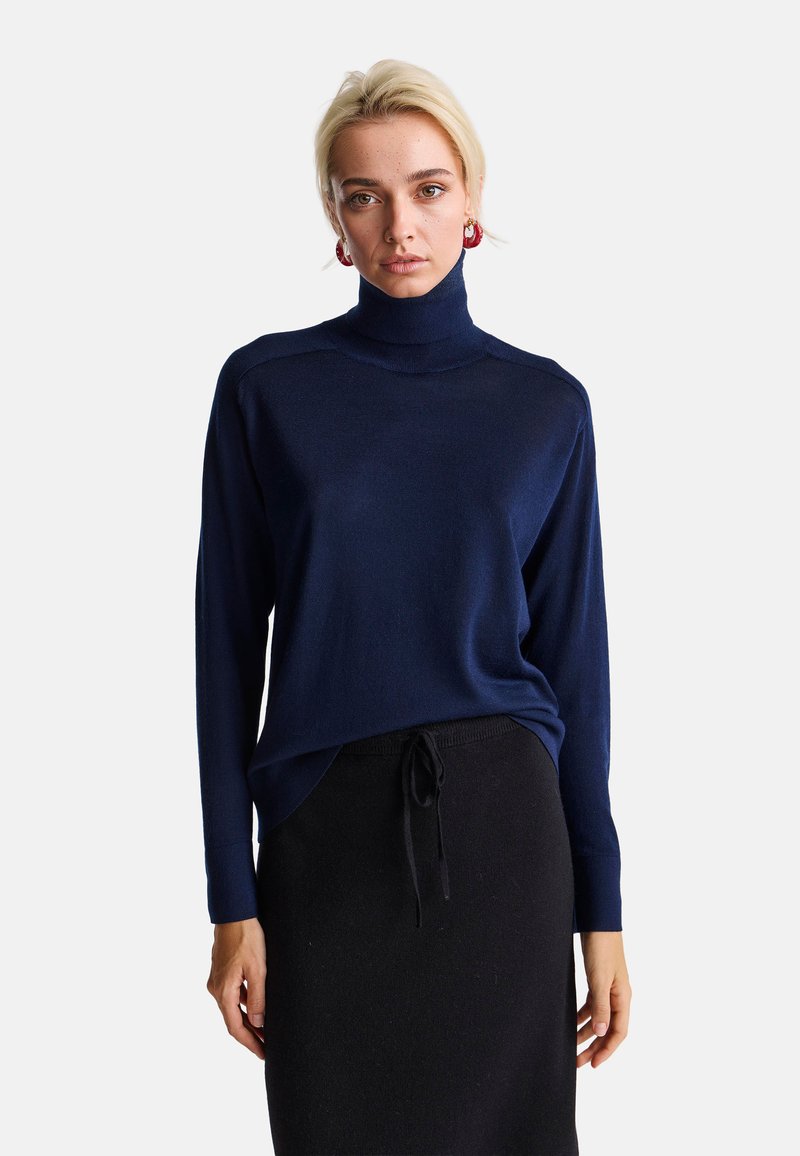 Maglione a collo alto blu navy con maniche lunghe, tessuto morbido e vestibilità rilassata, abbinato a una gonna nera con vita regolabile.