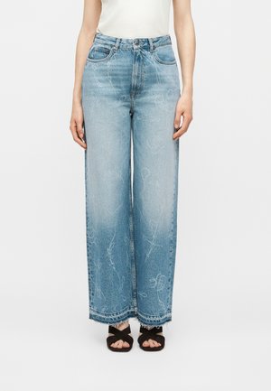 Femme portant un jean taille haute, jambes larges, bleu clair avec des motifs abstraits blancs délavés et des sandales noires croisées.