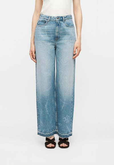 Mujer con vaqueros azul claro de tiro alto y pierna ancha con patrones abstractos blancos descoloridos y sandalias negras con tiras cruzadas.