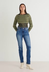 Suéter de punto corto en color caqui con patrón de tejido abierto, superpuesto sobre una blusa verde oscura. Unos jeans rectos azules y botas de tobillo beige completan el atuendo.