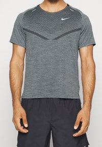 Harmaa lyhythihainen urheilupaita, jossa on teksturoitu kuvio, pyöreä pääntie ja heijastavat yksityiskohdat, sekä pieni Nike-logo rinnassa.