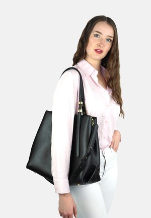 Zwarte leren tote bag met gestructureerde vorm, voorzien van gouden hardware, een zijritszak en dubbele handvatten. Draagt boven een roze shirt.