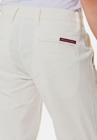 Pantalon en coton blanc avec une poche arrière munie d'une étiquette bordeaux. Le design comprend une coupe droite et des détails de couture visibles.