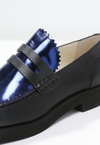 Mocassin en cuir noir avec dessus métallique bleu brillant présentant un bord en zigzag et un détail de bride noire.
