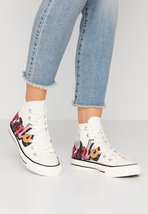 Witte hoge sneakers met graffiti-stijl letters in zwart, roze en oranje, gedragen met lichtblauwe jeans met rafelige zoom op een lichte achtergrond.