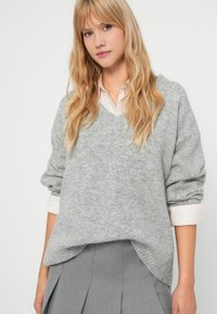 Szary sweter robiony na drutach z dekoltem w serek, luźny krój i podwinięte rękawy, nałożony na jasnoróżową koszulę z kołnierzykiem, zestawiony z szarą spódnicą plisowaną.