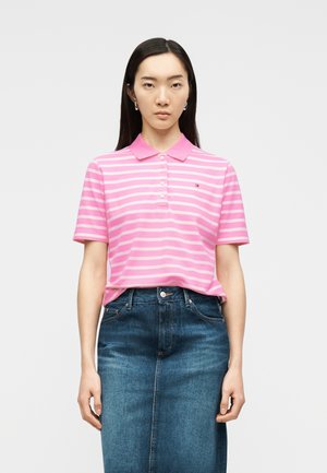 Mlada ženska v roza in beli polo majici s črtami, združeni z visokim modrim jeansi krilom, stoji pred enobarvnim ozadjem.