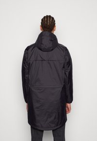 Veste noire à capuche imperméable avec des manches longues, une poche arrière zippée et une texture lisse. Présente un ourlet incurvé au bas.