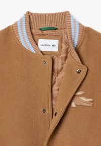 Giacca in pile marrone con colletto a coste decorato con strisce azzurro chiaro. Presenta chiusure a pressione, fodera trapuntata e un accento del logo beige.