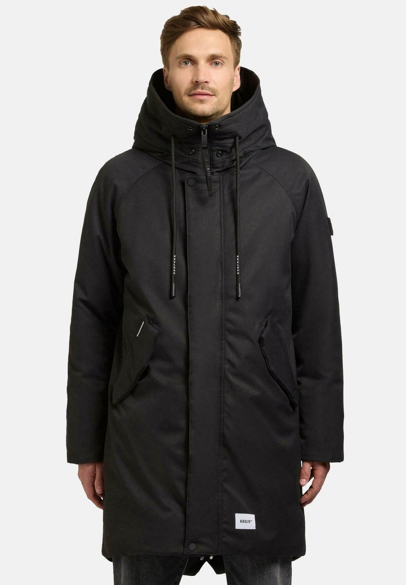 Sort parka-jakke med en stor hætte, der har lynlås og trykknaplukninger, sidelommer og subtil branding i kanten. Glat stoffinish.