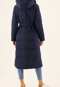 Abrigo de plumas navy con capucha, cintura ceñida y diseño acolchado. Presenta mangas largas y longitud hasta la mitad de la pantorrilla, combinado con jeans claros y zapatos blancos.