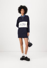 Mornarska haljina-sweatshirt s plavim traper ovratnikom, s bijelim panelom i natpisom "New Haven." Crne platforme i torba od tkanine.