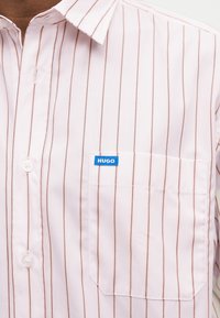 Λευκό πουκάμισο με κουμπιά, με λεπτές κάθετες καφέ ρίγες, τσέπη στο στήθος και μια μικρή μπλε ετικέτα με την επιγραφή "HUGO" στην τσέπη.