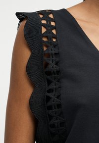 Μαύρο τοπ χωρίς μανίκια με κυματιστό κεντητό δαντελένιο τελείωμα κατά μήκος του ώμου και του άνω βραχίονα.