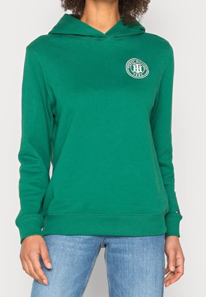 Personne portant un sweat à capuche vert Tommy Hilfiger avec logo sur la poitrine et la manche, associé à un jean bleu clair, debout devant un fond uni.