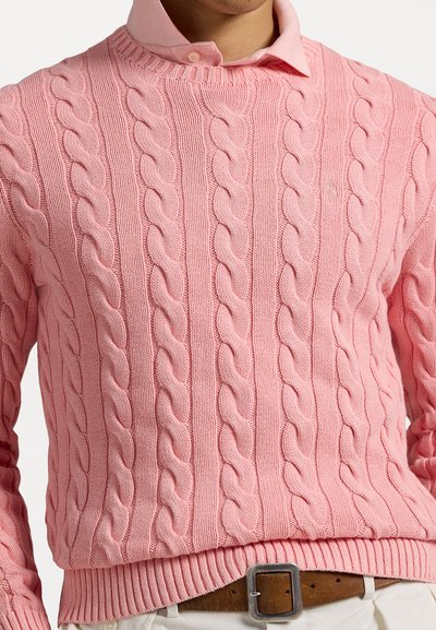 Różowy sweter z splotem kablowym, z teksturowanym wzorem, żebrowanym kołnierzem i dołem, noszony na jasnoróżowej koszuli z kołnierzykiem.