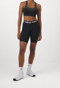 Brassière de sport noire avec tissu texturé, associée à un short noir taille haute avec une ceinture "NIKE PRO". Chaussures de sport blanches et chaussettes.