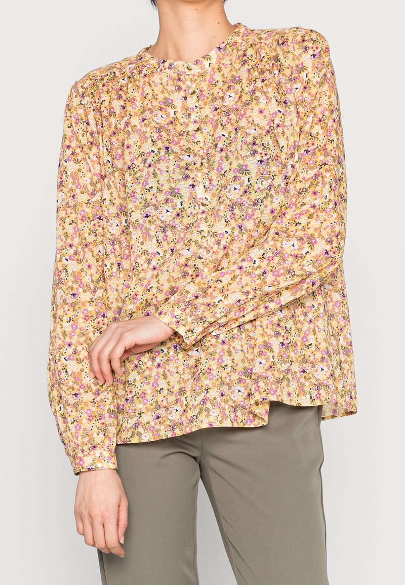 Blusa floreale gialla con motivi viola e rosa, maniche lunghe, scollatura rotonda e dettagli con bottoni. Tessuto morbido con vestibilità comoda.