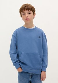 Felpa blu realizzata in tessuto morbido, con una tasca frontale, polsini a coste e un piccolo logo di teschio nero sul petto. Indossata con jeans.
