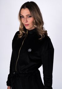 Naine laineliste helepruunide juustega, seljas must lukuga dressipluus, millel on rinnal väike valge tikitud logo, seisab ühevärvilise tausta ees.