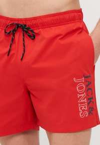 Piros fürdőnadrág könnyű anyagból, gumis derékkal és fekete húzózsinórral. Fehér és kék "JACK & JONES" logóval van ellátva.