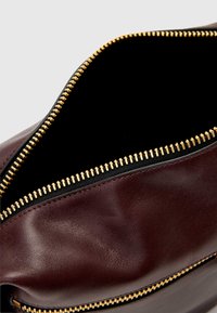 Mala de couro marrom com um zíper dourado. O interior é preto, apresentando uma textura suave e uma forma estruturada. Detalhe do zíper claramente visível.