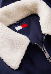 Μπουφάν ναυτικό μπλε με λευκή γιακά sherpa fleece και λογότυπο Tommy Jeans στην εσωτερική ετικέτα κοντά στο μεταλλικό φερμουάρ.