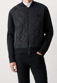 Veste bomber noire avec devant en nylon matelassé, manches en tricot côtelé et fermeture éclair. Détail logo simple sur la poitrine, finition mate.