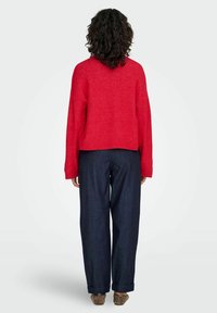 Pull rouge en maille avec une coupe ample, des épaules tombantes et des poignets côtelés, associé à un pantalon en denim bleu foncé à jambes larges.