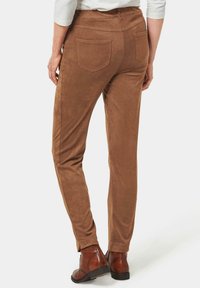 Pantalon en daim marron avec une coupe slim, comportant des poches arrière et une texture lisse. Porté avec des bottines brunes pour un look harmonieux.