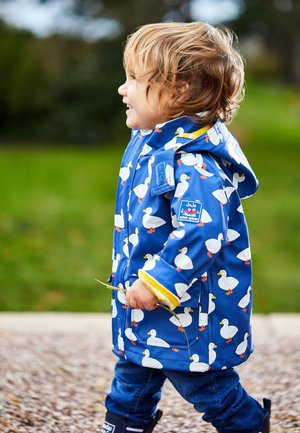 Blauer Regenmantel mit weißen Entenmustern, gelben Akzenten und einer Kapuze. Das Kind trägt außerdem blaue Jeans und Gummistiefel.