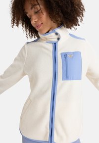 Giacca in pile color crema con accenti azzurri chiari. Presenta un colletto alto, chiusura con zip e una tasca sul petto. Tessuto strutturato per il calore.