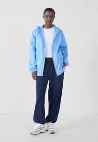 Chaqueta azul claro, resistente al agua, con puños elásticos y cremallera frontal; combinada con pantalones navy con cordón y zapatillas deportivas plateadas.