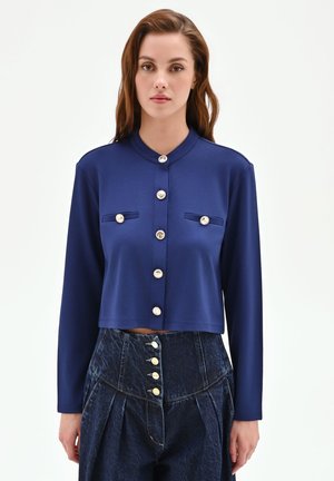 Donna che indossa una giacca corta blu navy a maniche lunghe con bottoni in perla e pantaloni di denim scuro a vita alta con più bottoni.